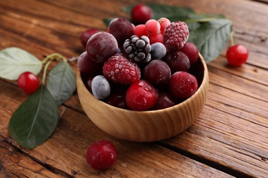 Photo of Mix of ripe frozen berries in bowl and green leaves on wooden table, closeup