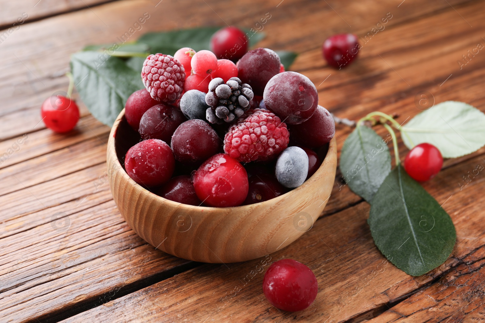 Photo of Mix of ripe frozen berries in bowl and green leaves on wooden table, closeup