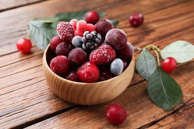 Photo of Mix of ripe frozen berries in bowl and green leaves on wooden table, closeup