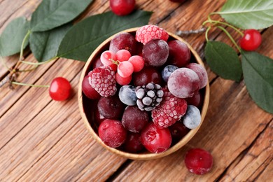 Photo of Mix of ripe frozen berries in bowl and green leaves on wooden table, flat lay