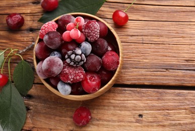 Photo of Mix of ripe frozen berries in bowl and green leaves on wooden table, flat lay. Space for text