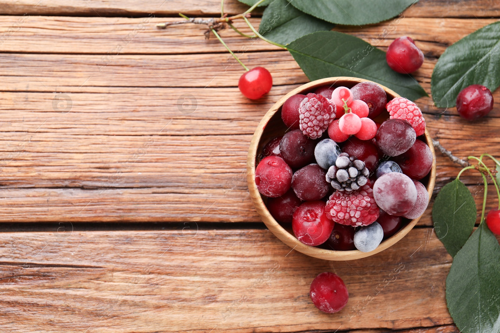 Photo of Mix of ripe frozen berries in bowl and green leaves on wooden table, flat lay. Space for text