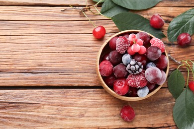 Photo of Mix of ripe frozen berries in bowl and green leaves on wooden table, flat lay. Space for text