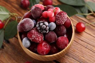 Photo of Mix of ripe frozen berries in bowl and green leaves on wooden table, closeup
