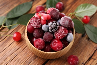 Photo of Mix of ripe frozen berries in bowl and green leaves on wooden table, closeup