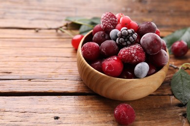 Photo of Mix of ripe frozen berries in bowl and green leaves on wooden table, closeup. Space for text