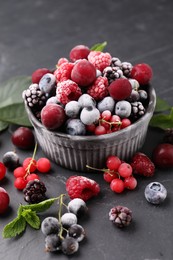 Photo of Mix of ripe frozen berries, leaves and bowl on dark textured table, closeup