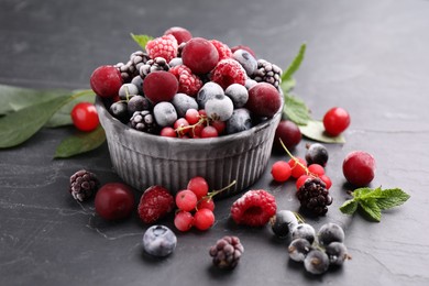 Photo of Mix of ripe frozen berries, leaves and bowl on dark textured table, closeup