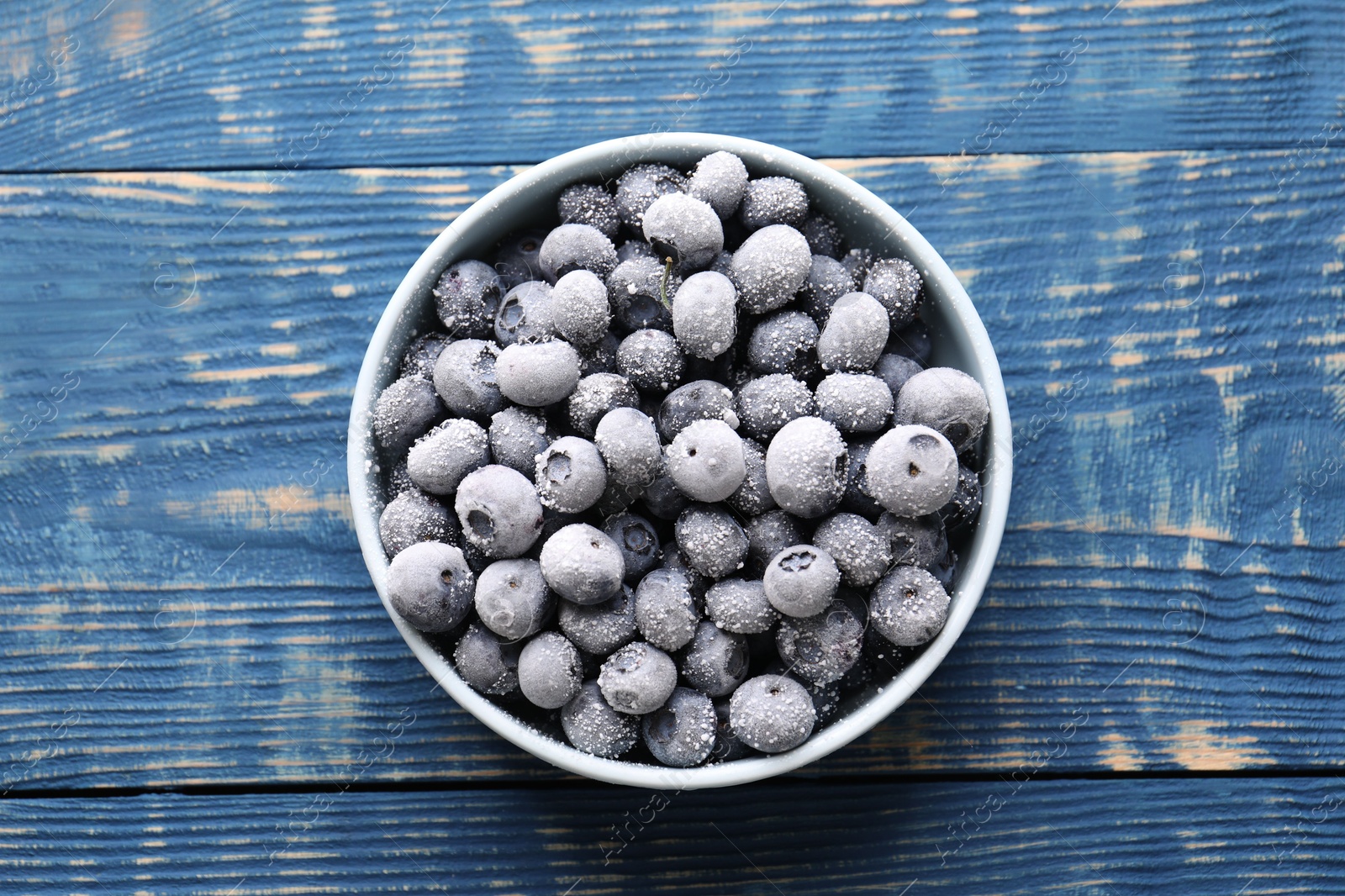 Photo of Ripe frozen blueberries in bowl on blue wooden table, top view