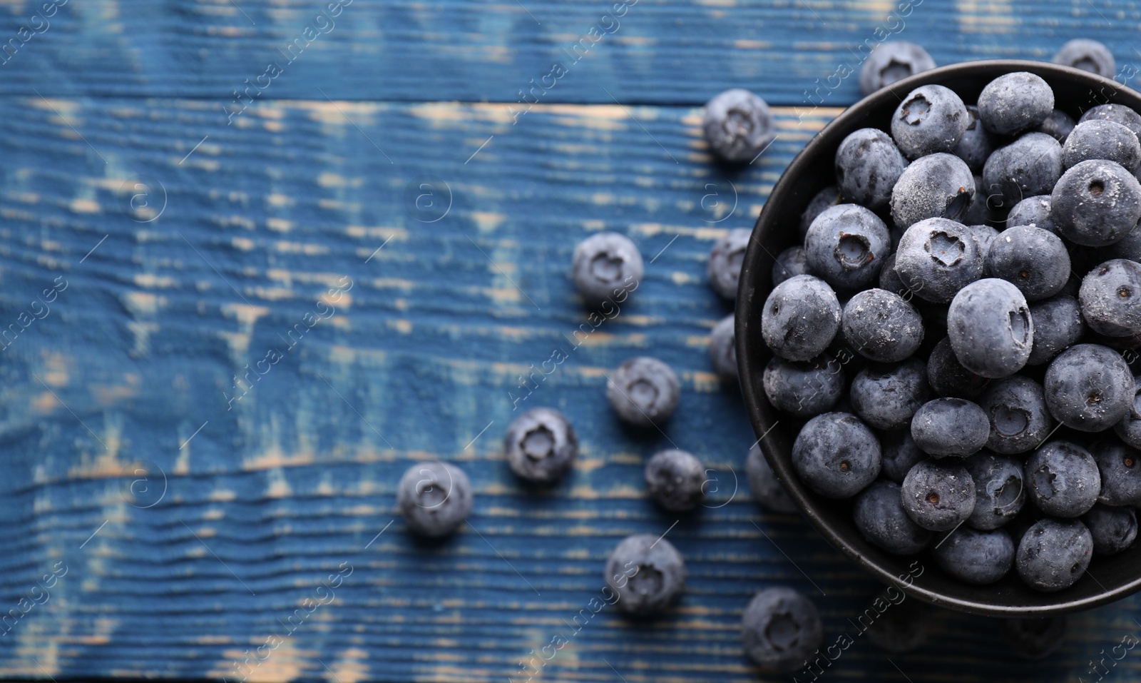 Photo of Ripe frozen blueberries in bowl on blue wooden table, flat lay. Space for text