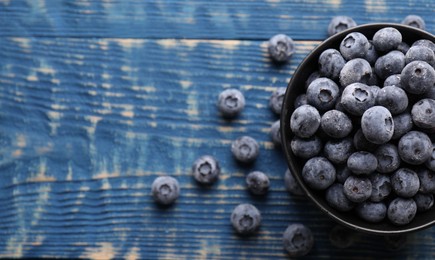 Photo of Ripe frozen blueberries in bowl on blue wooden table, flat lay. Space for text
