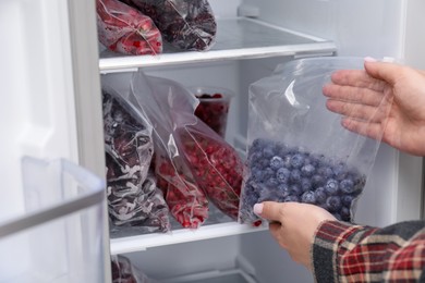 Photo of Freezing berries. Woman putting plastic bag with blueberries into open fridge, closeup
