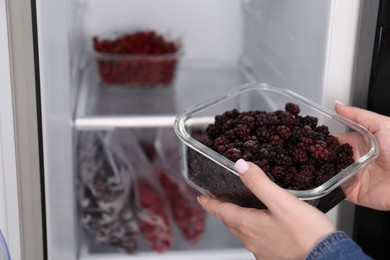 Photo of Freezing berries. Woman putting container with blackberries into open fridge, closeup