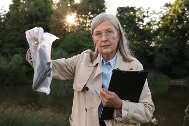 Photo of Forensic expertise. Detective with clipboard and evidence at crime scene near lake outdoors