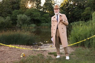 Photo of Forensic expertise. Detective with smoking pipe and clipboard working at crime scene near lake outdoors, space for text