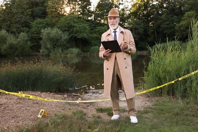 Photo of Forensic expertise. Detective with smoking pipe and clipboard working at crime scene near lake outdoors, space for text