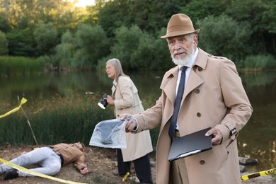 Photo of Forensic expertise. Detectives working at crime scene with dead man's body near lake outdoors