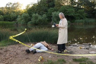 Photo of Forensic expertise. Detective with camera working at crime scene with dead man's body near lake outdoors