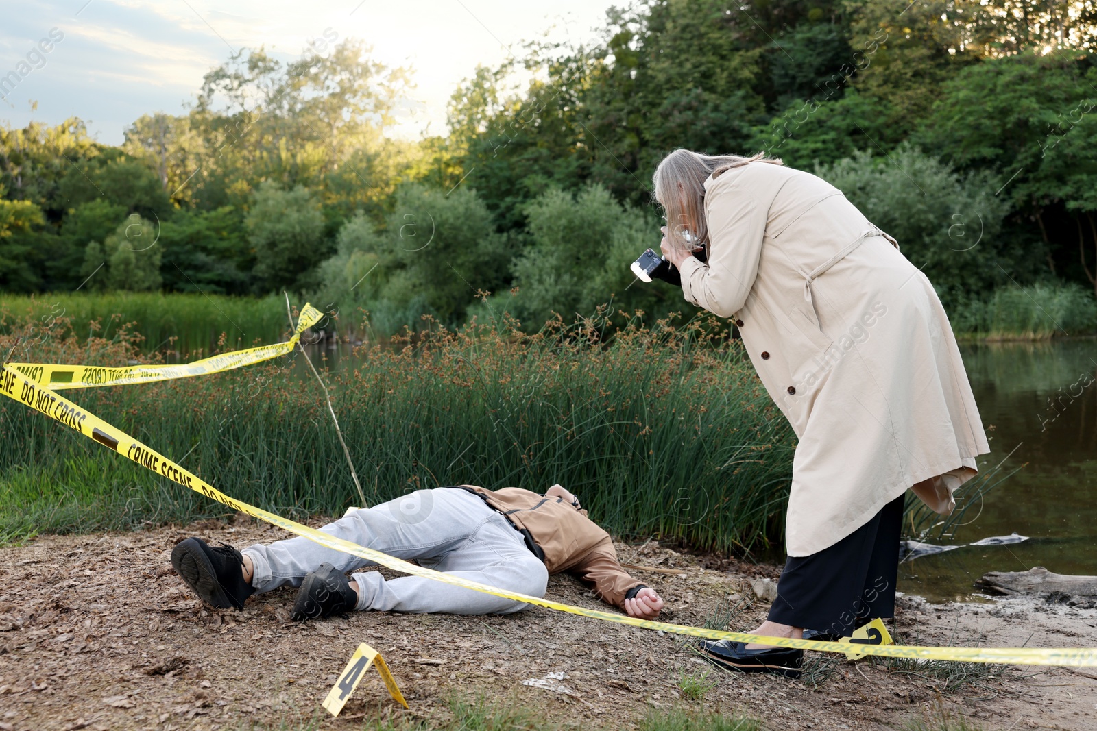 Photo of Forensic expertise. Detective with camera working at crime scene with dead man's body near lake outdoors