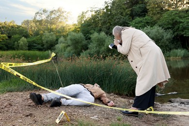 Photo of Forensic expertise. Detective with camera working at crime scene with dead man's body near lake outdoors