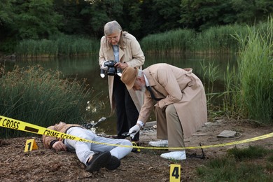 Photo of Forensic expertise. Detectives working at crime scene with dead man's body near lake outdoors
