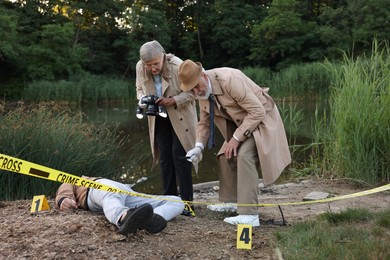 Photo of Forensic expertise. Detectives working at crime scene with dead man's body near lake outdoors