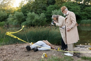 Photo of Forensic expertise. Detectives working at crime scene with dead man's body near lake outdoors