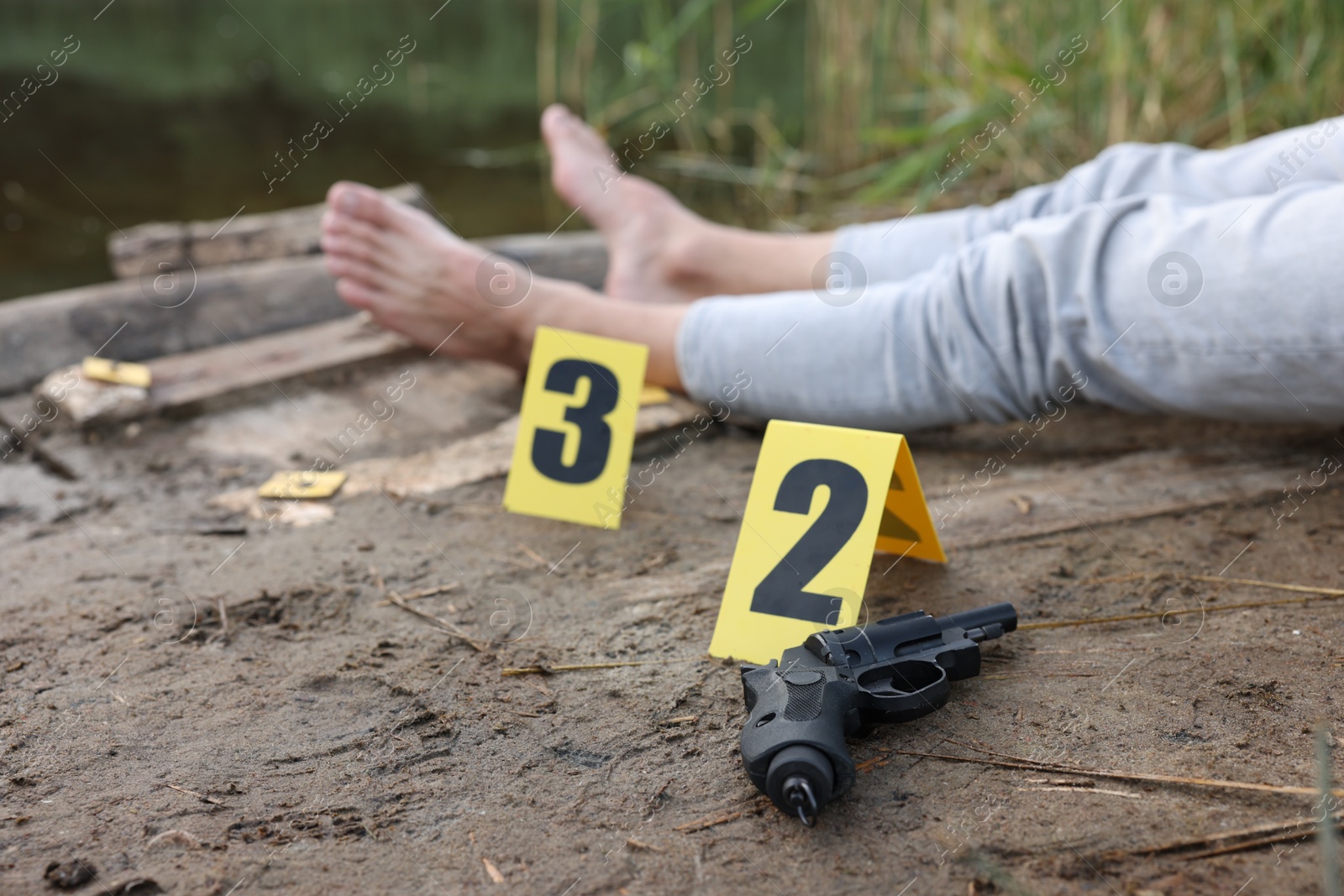 Photo of Forensic expertise. Crime scene with dead man's body, markers and gun near lake outdoors, selective focus