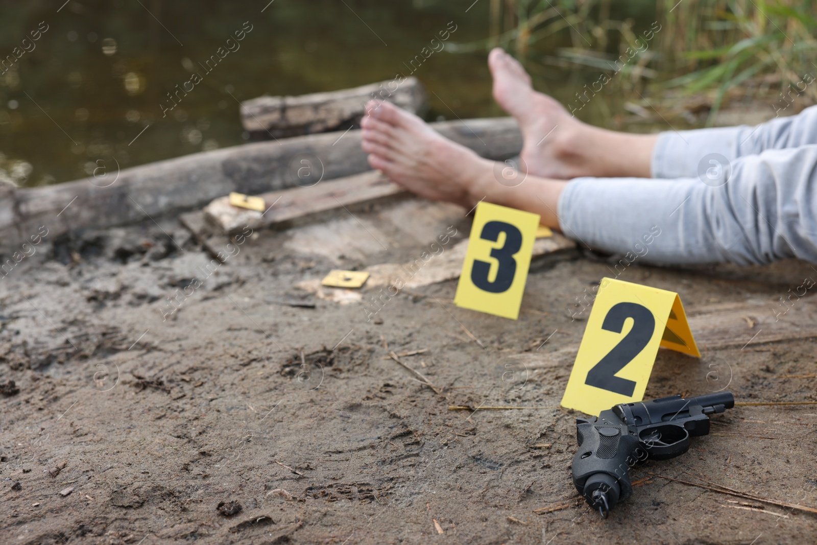 Photo of Forensic expertise. Crime scene with dead man's body, markers and gun near lake outdoors, selective focus. Space for text