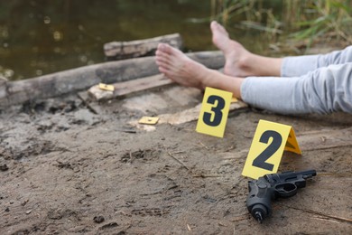 Photo of Forensic expertise. Crime scene with dead man's body, markers and gun near lake outdoors, selective focus. Space for text