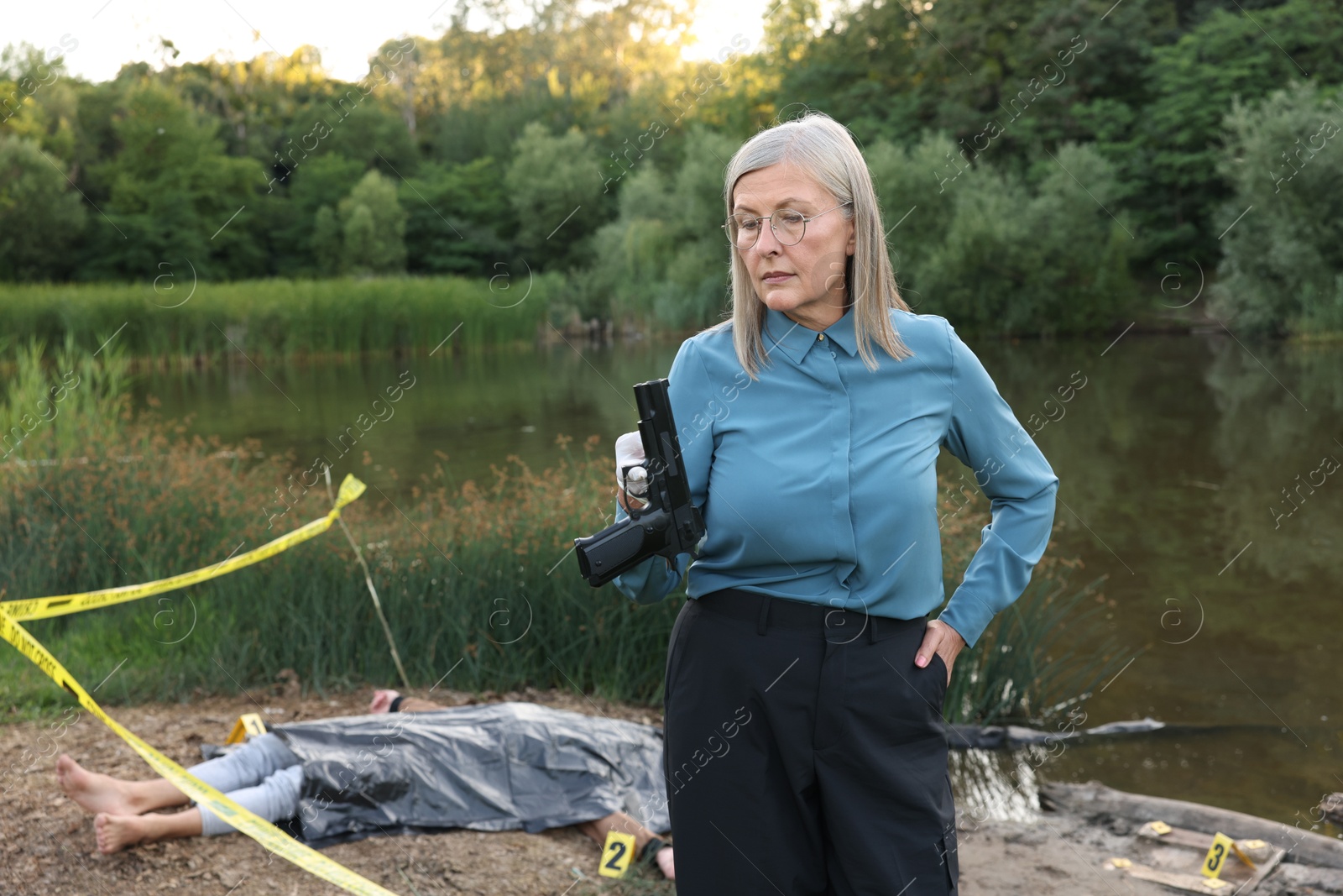 Photo of Forensic expertise. Detective holding evidence working at crime scene with dead man's body near lake outdoors