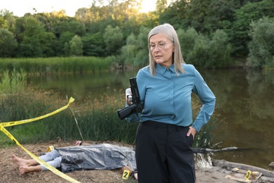 Photo of Forensic expertise. Detective holding evidence working at crime scene with dead man's body near lake outdoors
