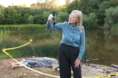 Photo of Forensic expertise. Detective holding evidence working at crime scene with dead man's body near lake outdoors