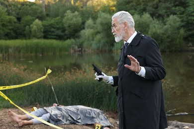 Photo of Forensic expertise. Detective with evidence working at crime scene with dead man's body near lake outdoors