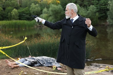 Photo of Forensic expertise. Detective with evidence working at crime scene with dead man's body near lake outdoors