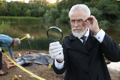 Photo of Forensic expertise. Detectives working at crime scene with dead man's body near lake outdoors, selective focus