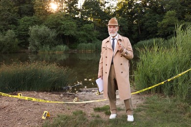 Photo of Forensic expertise. Detective with smoking pipe and clipboard working at crime scene near lake outdoors, space for text