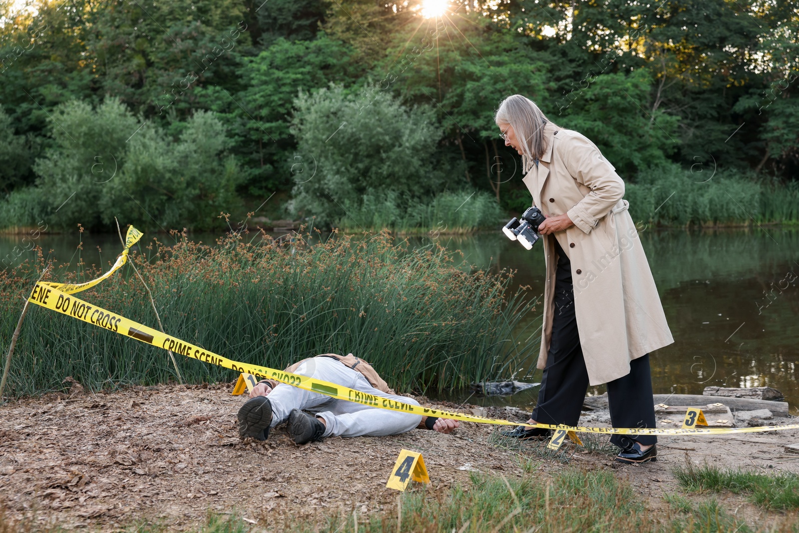 Photo of Forensic expertise. Detective with camera working at crime scene with dead man's body near lake outdoors