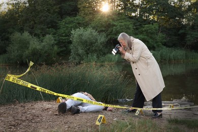 Photo of Forensic expertise. Detective with camera working at crime scene with dead man's body near lake outdoors