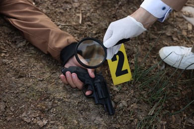 Photo of Forensic expertise. Detective with magnifying glass working at crime scene with dead man's body outdoors, closeup