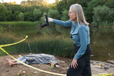 Photo of Forensic expertise. Detective holding evidence working at crime scene with dead man's body near lake outdoors