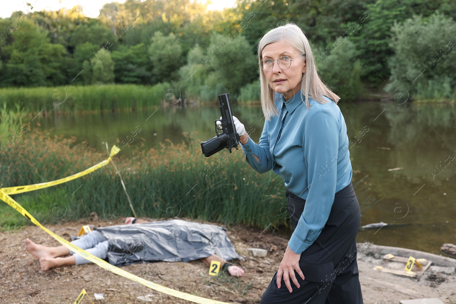 Photo of Forensic expertise. Detective holding evidence working at crime scene with dead man's body near lake outdoors