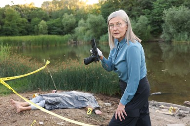 Photo of Forensic expertise. Detective holding evidence working at crime scene with dead man's body near lake outdoors