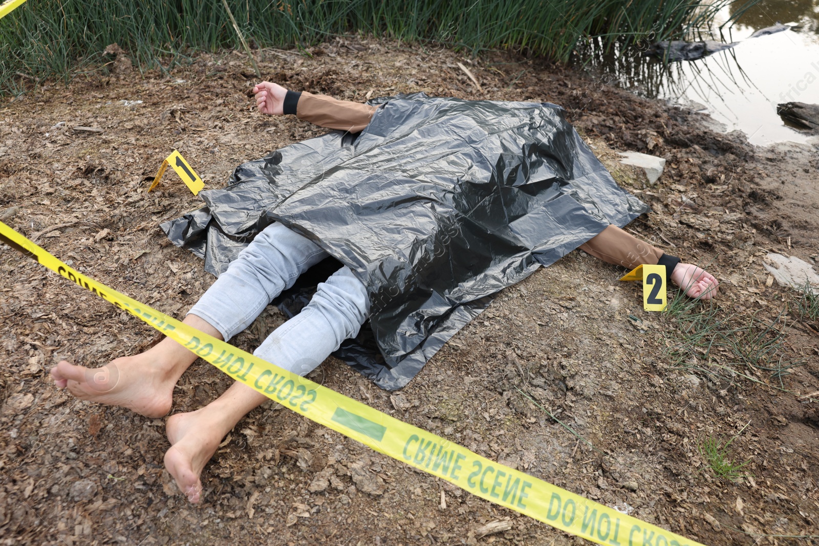 Photo of Forensic expertise. Crime scene with dead man's body, markers and police tape near lake outdoors