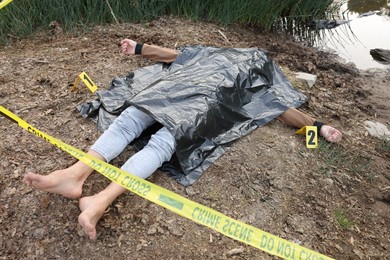 Photo of Forensic expertise. Crime scene with dead man's body, markers and police tape near lake outdoors