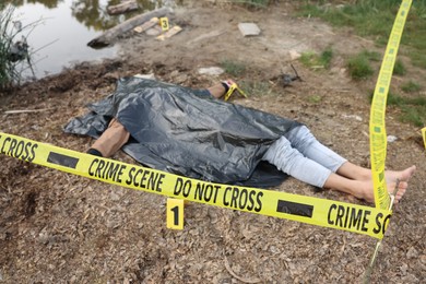 Photo of Forensic expertise. Crime scene with dead man's body, markers and police tape near lake outdoors, selective focus