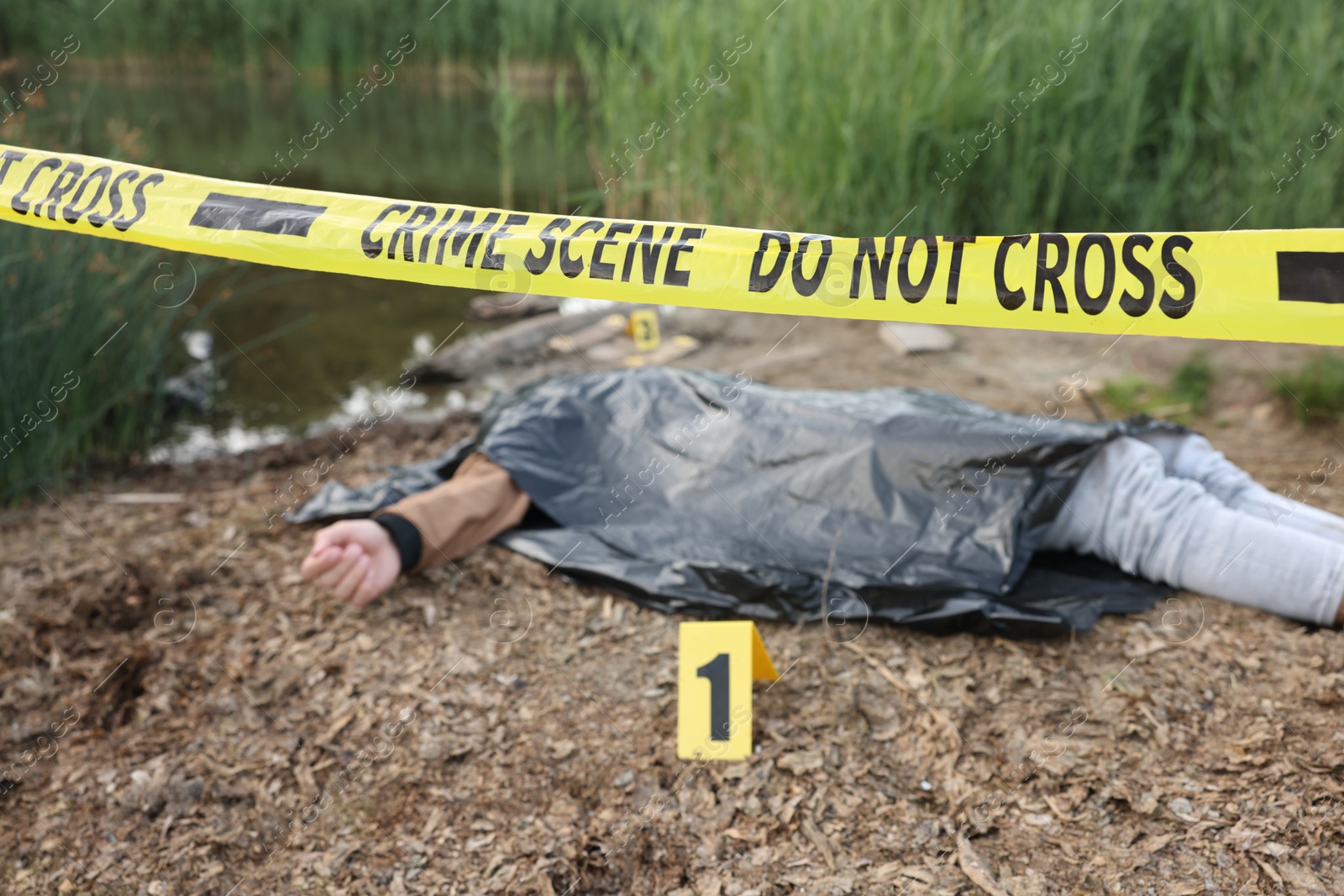 Photo of Forensic expertise. Crime scene with dead man's body, marker and police tape near lake outdoors, selective focus