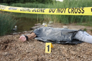 Photo of Forensic expertise. Crime scene with dead man's body, marker and police tape near lake outdoors, selective focus