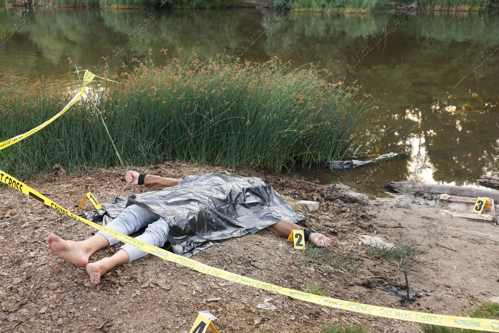 Photo of Forensic expertise. Crime scene with dead man's body, markers and police tape near lake outdoors