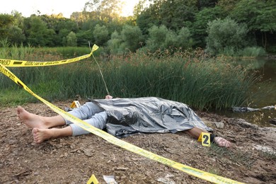 Photo of Forensic expertise. Crime scene with dead man's body, markers and police tape near lake outdoors